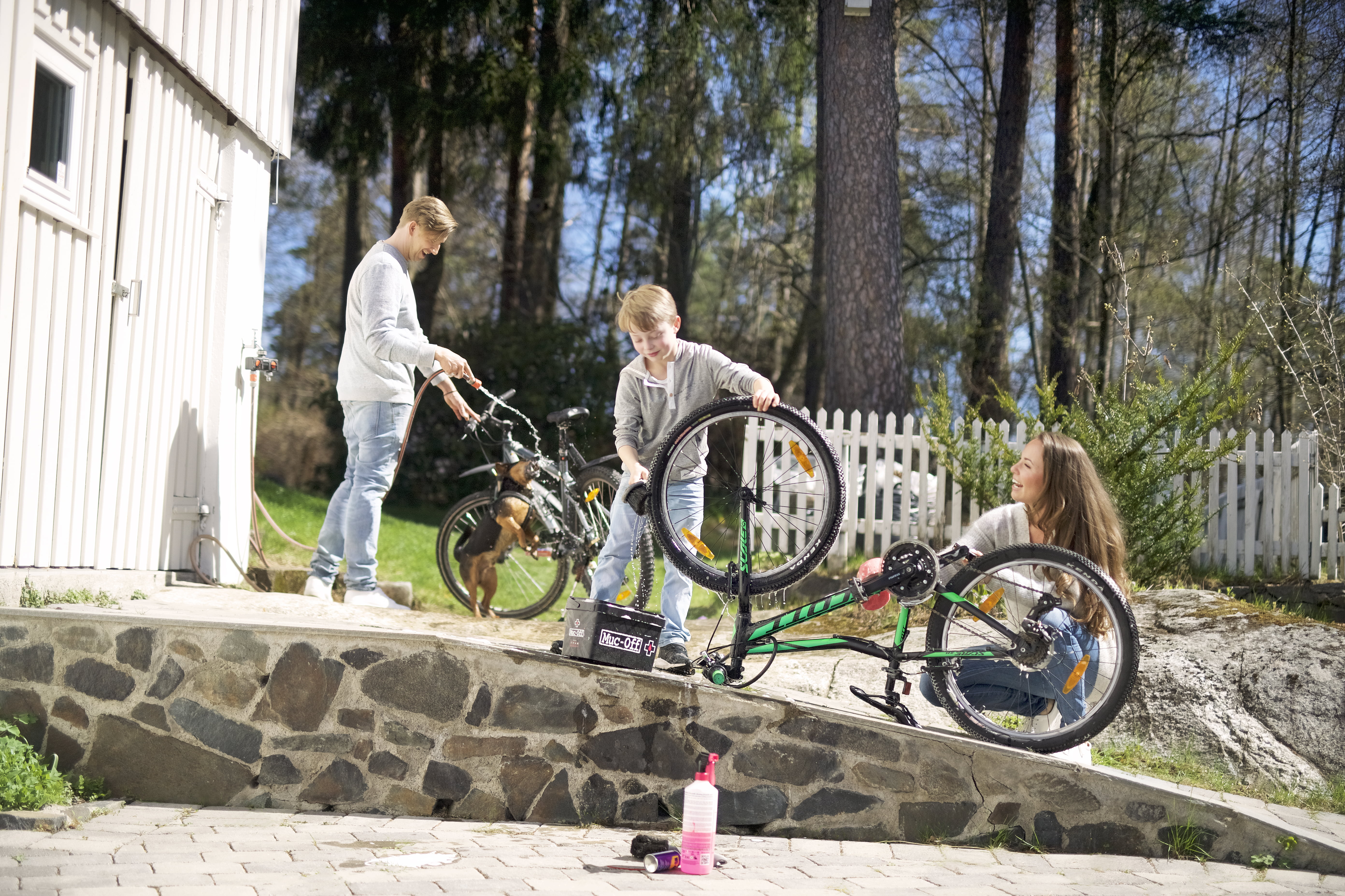 Tre personer og en hund ute i hagen reparerer sykler ved en steinmur, med verktøykasse og drikkeflaske på bakken.