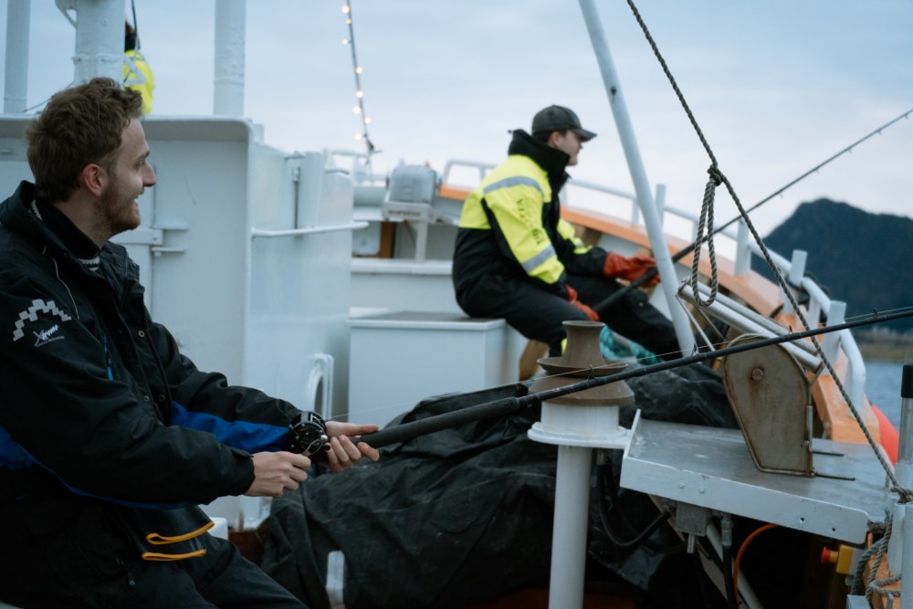 To personer fisker fra en båt med fjell i bakgrunnen.